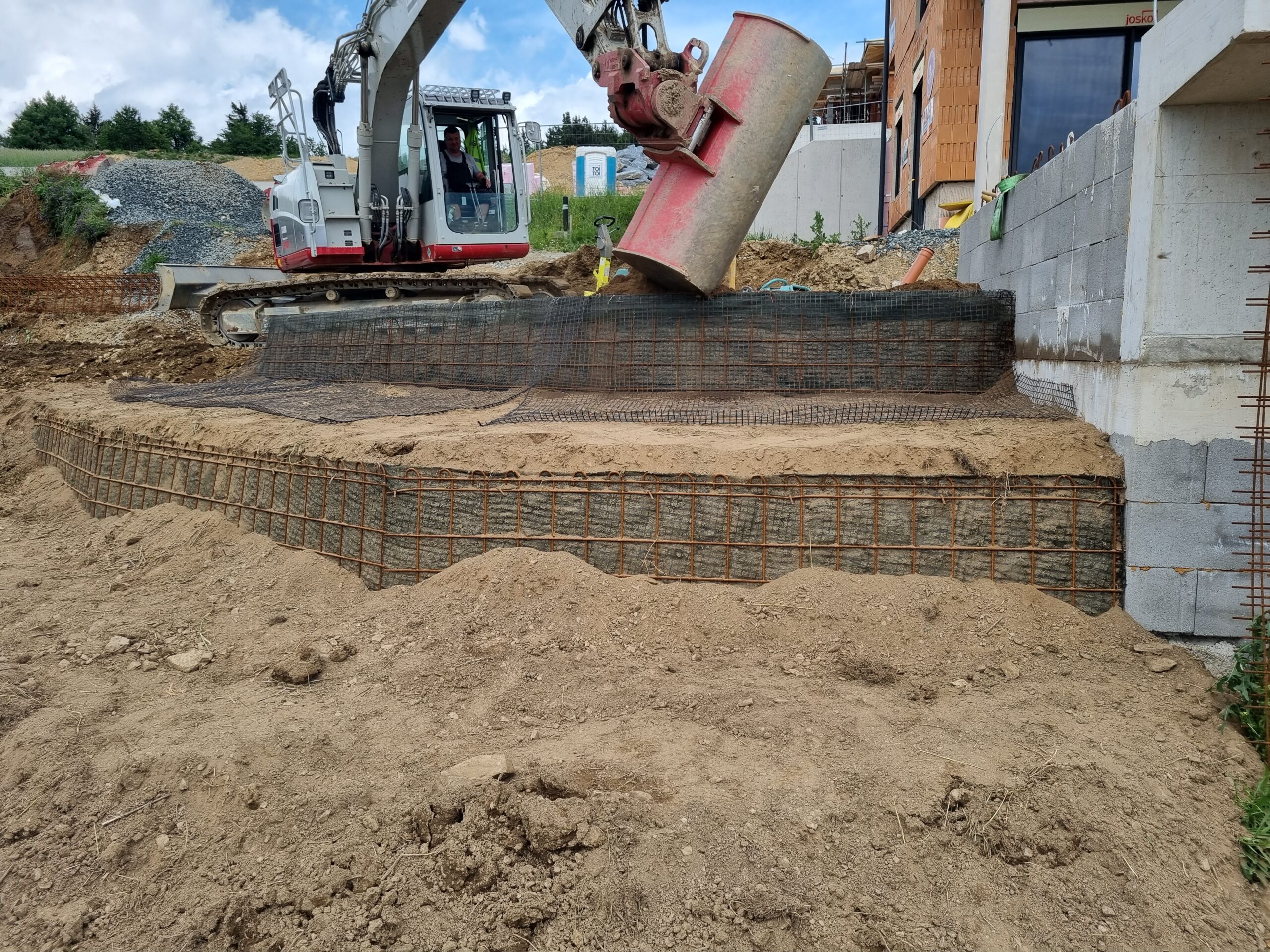 Bagger steht neben einem Rohbau und positioniert ein Anbaugerät; im Vordergrund ist eine verschalte und bewehrte Fundamentstufe mit Stahlmatten und Bewehrungskorb im sandigen Aushub zu sehen.