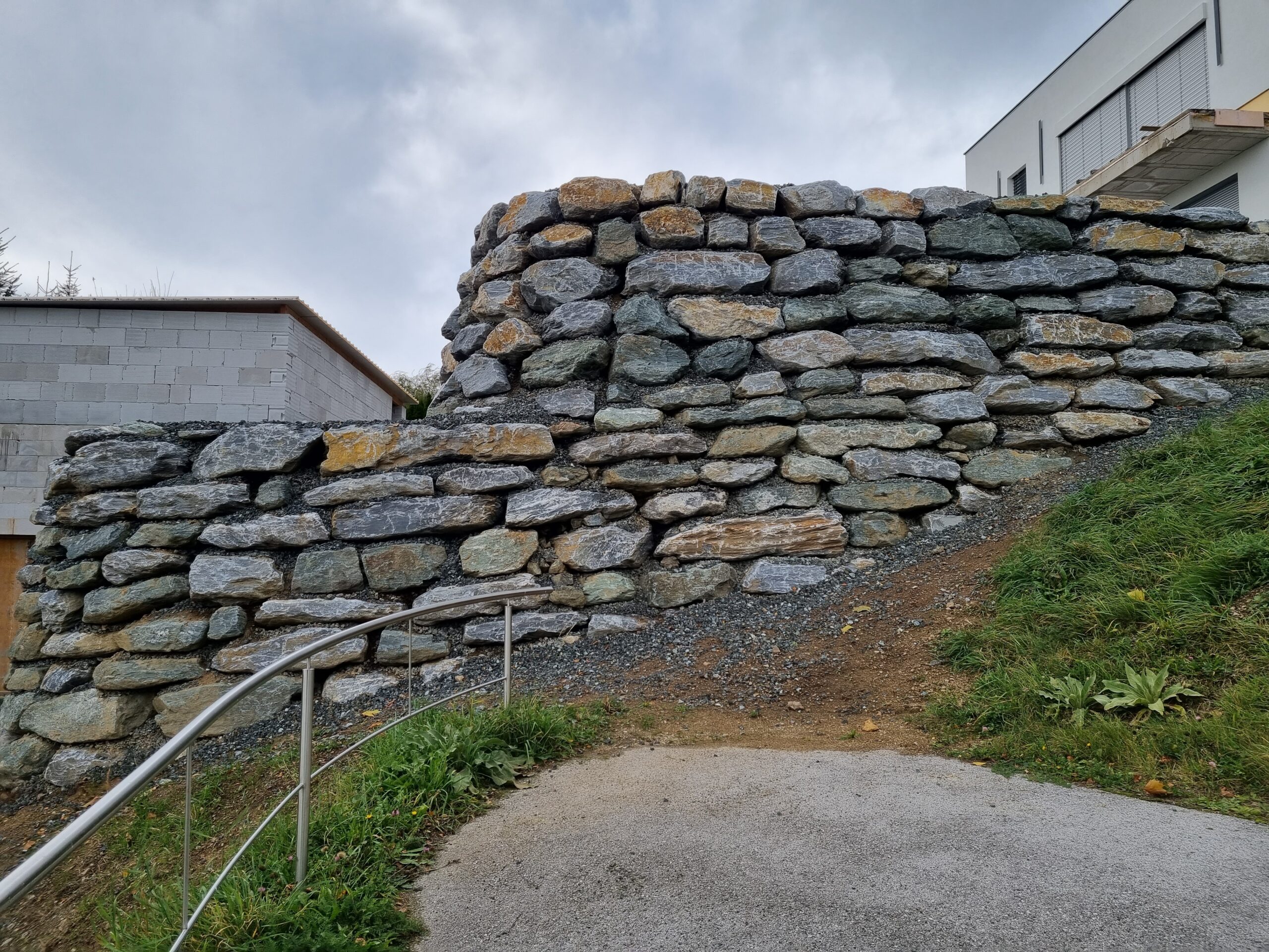 Große Naturstein-Stützmauer am Hang neben einem Weg mit Geländer.