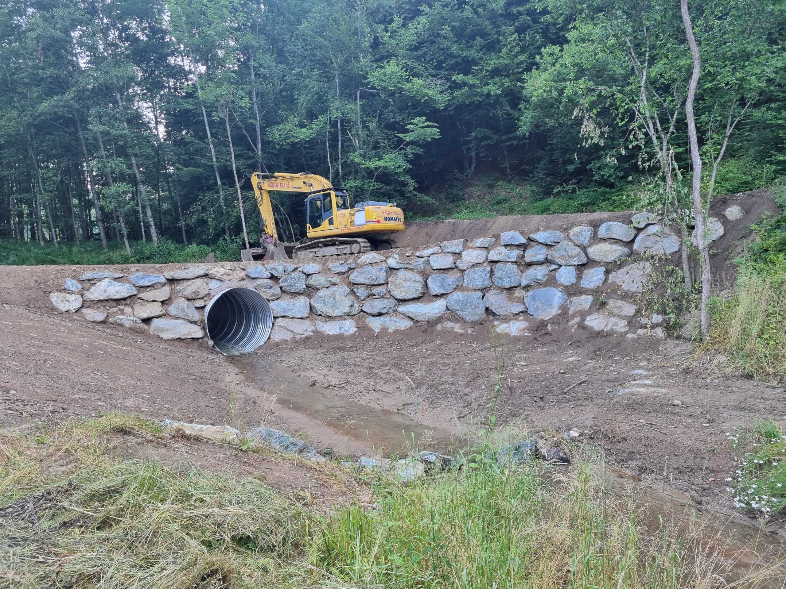 Ein gelber Bagger steht auf einer Natursteinbrücke und saniert einen Bachlauf. 